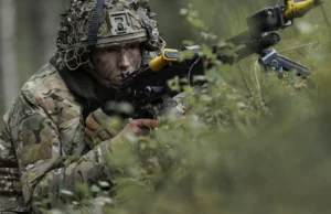 British troops test drone war tactics near Russian border Soldier in camouflage aiming a rifle while crouched among dense forest vegetation.