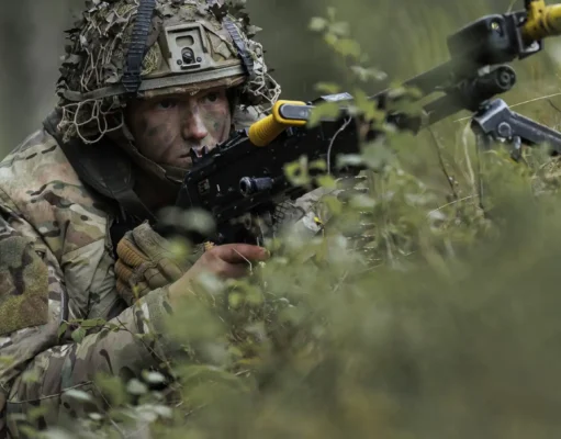British troops test drone war tactics near Russian border Soldier in camouflage aiming a rifle while crouched among dense forest vegetation.