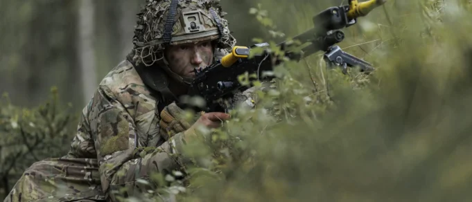 Soldier in camouflage aiming a rifle while crouched among dense forest vegetation.