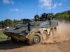 Wheeled armored vehicle driving along a dirt road, kicking up a cloud of dust with crew visible in the open hatchs atop the vehicle.
