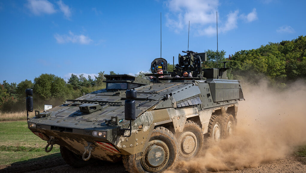 Wheeled armored vehicle driving along a dirt road, kicking up a cloud of dust with crew visible in the open hatchs atop the vehicle.