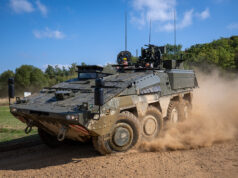 Wheeled armored vehicle driving along a dirt road, kicking up a cloud of dust with crew visible in the open hatchs atop the vehicle.