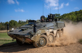 Wheeled armored vehicle driving along a dirt road, kicking up a cloud of dust with crew visible in the open hatchs atop the vehicle.
