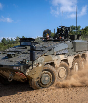 All Boxers built to one standard, says minister Wheeled armored vehicle driving along a dirt road, kicking up a cloud of dust with crew visible in the open hatchs atop the vehicle.