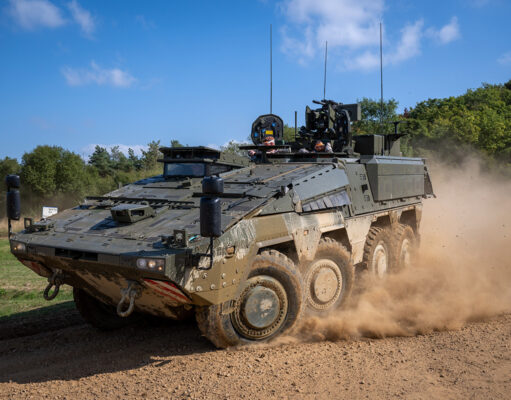 Wheeled armored vehicle driving along a dirt road, kicking up a cloud of dust with crew visible in the open hatchs atop the vehicle.