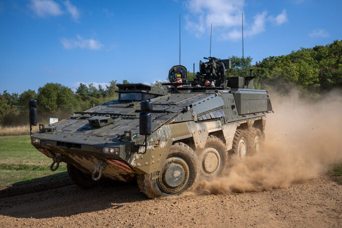 Wheeled armored vehicle driving along a dirt road, kicking up a cloud of dust with crew visible in the open hatchs atop the vehicle.