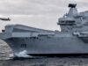 A large gray aircraft carrier cutting through rough seas, hull number RO8, with two small airborne objects (drones) flying nearby.