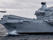 British aircraft carrier to fly combat drone within 18 months A large gray aircraft carrier cutting through rough seas, hull number RO8, with two small airborne objects (drones) flying nearby.