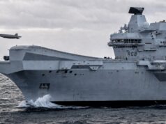British aircraft carrier to fly combat drone within 18 months A large gray aircraft carrier cutting through rough seas, hull number RO8, with two small airborne objects (drones) flying nearby.