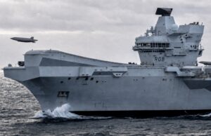 British aircraft carrier to fly combat drone within 18 months A large gray aircraft carrier cutting through rough seas, hull number RO8, with two small airborne objects (drones) flying nearby.