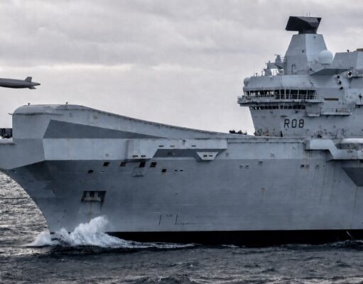 British aircraft carrier to fly combat drone within 18 months A large gray aircraft carrier cutting through rough seas, hull number RO8, with two small airborne objects (drones) flying nearby.