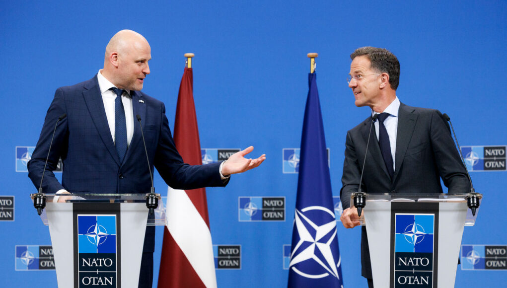 cq5dam.web.1280.1280 (15) Two suited men stand at podiums with NATO logos, speaking to each other during a press conference, with a Latvia flag and a NATO flag behind them.
