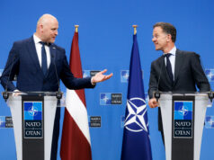 Two suited men stand at podiums with NATO logos, speaking to each other during a press conference, with a Latvia flag and a NATO flag behind them.