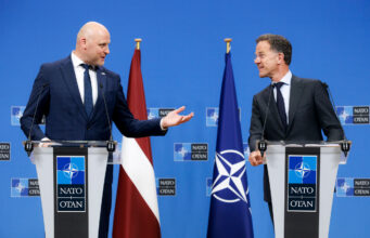 Two suited men stand at podiums with NATO logos, speaking to each other during a press conference, with a Latvia flag and a NATO flag behind them.