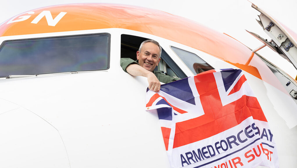 Smiling pilot leans from the cockpit of a white and orange airplane, holding a Union Jack flag that reads Armed Forces Day: Show Your Support.