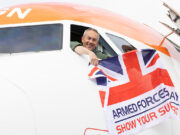 Smiling pilot leans from the cockpit of a white and orange airplane, holding a Union Jack flag that reads Armed Forces Day: Show Your Support.
