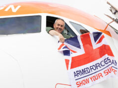 Smiling pilot leans from the cockpit of a white and orange airplane, holding a Union Jack flag that reads Armed Forces Day: Show Your Support.