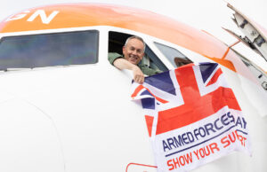 Smiling pilot leans from the cockpit of a white and orange airplane, holding a Union Jack flag that reads Armed Forces Day: Show Your Support.