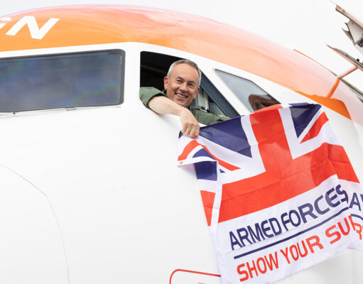 Smiling pilot leans from the cockpit of a white and orange airplane, holding a Union Jack flag that reads Armed Forces Day: Show Your Support.