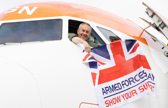 Smiling pilot leans from the cockpit of a white and orange airplane, holding a Union Jack flag that reads Armed Forces Day: Show Your Support.