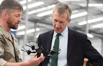 New Defence Secretary opens huge drone centre in Swindon Military personnel in camouflage hands a tactical vest to a businessman in a suit inside a hangar, as they converse.