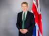 Portrait of a man in a dark suit and green tie standing beside a Union Jack flag behind him. (informative)