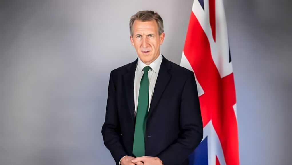 enhanced Portrait of a man in a dark suit and green tie standing beside a Union Jack flag behind him. (informative)