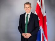 Portrait of a man in a dark suit and green tie standing beside a Union Jack flag behind him. (informative)