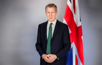 Portrait of a man in a dark suit and green tie standing beside a Union Jack flag behind him. (informative)