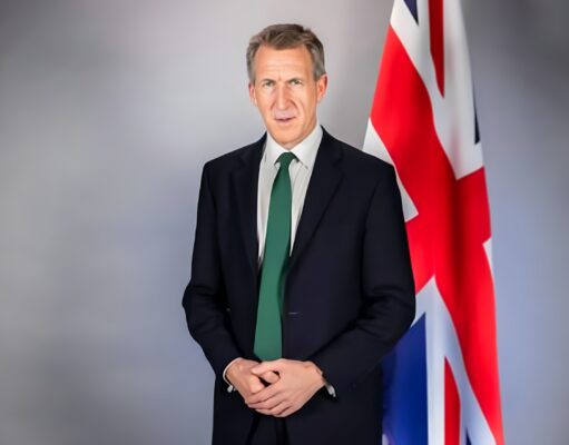 Portrait of a man in a dark suit and green tie standing beside a Union Jack flag behind him. (informative)