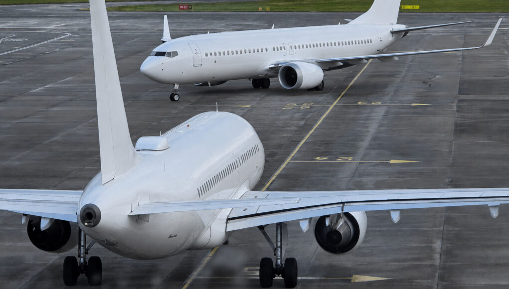 Two large white commercial aircraft on an airport tarmac, one in the foreground viewed from the rear with engines and tail visible, another taxiing in the background.