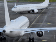 Two large white commercial aircraft on an airport tarmac, one in the foreground viewed from the rear with engines and tail visible, another taxiing in the background.