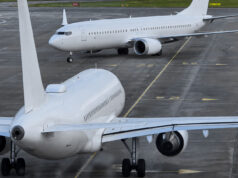 Two large white commercial aircraft on an airport tarmac, one in the foreground viewed from the rear with engines and tail visible, another taxiing in the background.