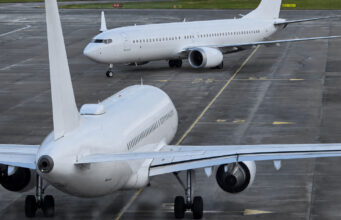 Two large white commercial aircraft on an airport tarmac, one in the foreground viewed from the rear with engines and tail visible, another taxiing in the background.