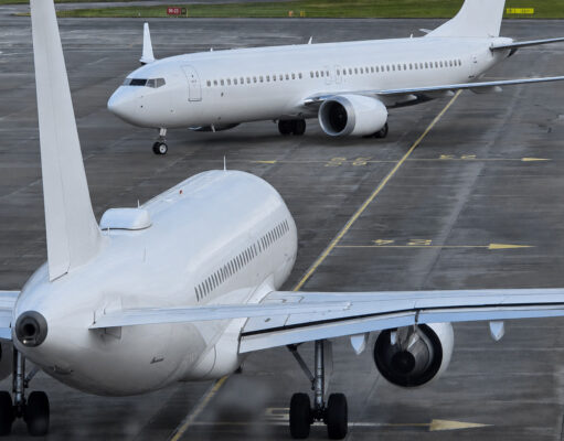 Two large white commercial aircraft on an airport tarmac, one in the foreground viewed from the rear with engines and tail visible, another taxiing in the background.