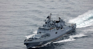 Overhead view of a gray military destroyer cutting through the ocean with the hull number 799 visible on the bow; wake behind the ship.