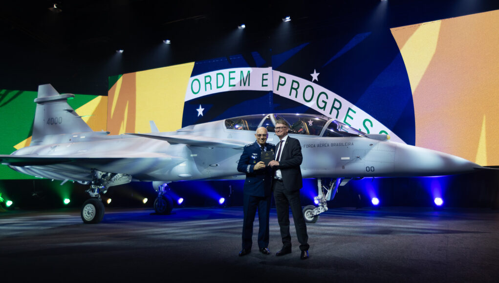 gripen-f Two men in formal attire stand beside a silver fighter jet on a stage, with a large Brazilian flag backdrop behind them and the cockpit visible to the right.