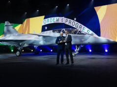 Saab rolls out first two-seat Gripen F for Brazil Two men in formal attire stand beside a silver fighter jet on a stage, with a large Brazilian flag backdrop behind them and the cockpit visible to the right.