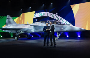 Two men in formal attire stand beside a silver fighter jet on a stage, with a large Brazilian flag backdrop behind them and the cockpit visible to the right.