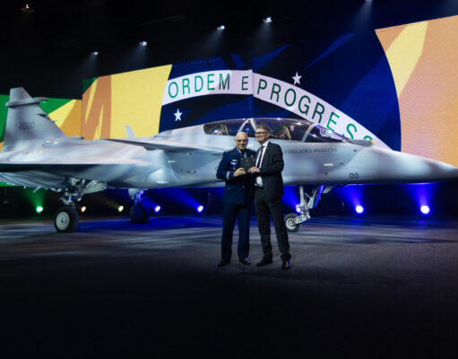 Saab rolls out first two-seat Gripen F for Brazil Two men in formal attire stand beside a silver fighter jet on a stage, with a large Brazilian flag backdrop behind them and the cockpit visible to the right.