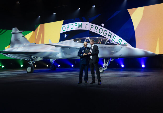 Two men in formal attire stand beside a silver fighter jet on a stage, with a large Brazilian flag backdrop behind them and the cockpit visible to the right.