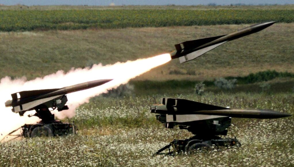 Two missiles launch from mobile ground launchers in a grassy field, producing bright flames and smoke trails.