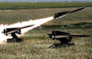 Two missiles launch from mobile ground launchers in a grassy field, producing bright flames and smoke trails.