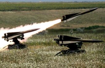US clears large Hawk sustainment package for Ukraine Two missiles launch from mobile ground launchers in a grassy field, producing bright flames and smoke trails.