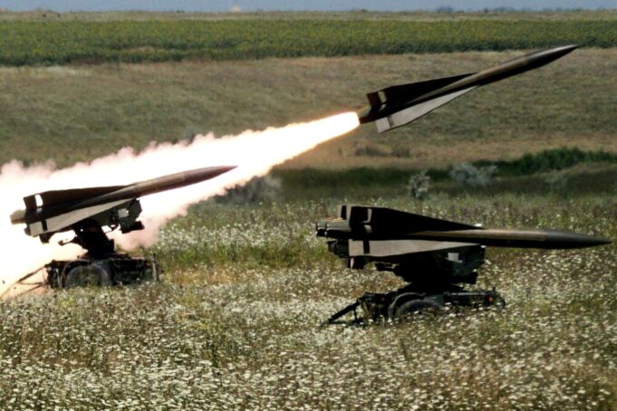 Two missiles launch from mobile ground launchers in a grassy field, producing bright flames and smoke trails.