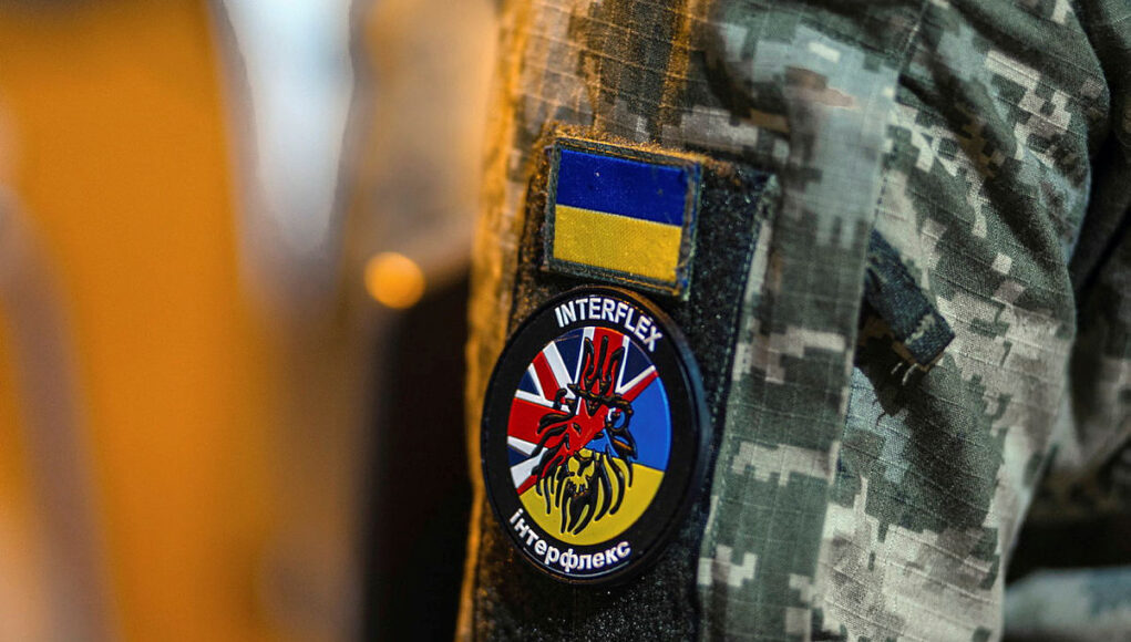 Military jacket with unit patches: Ukrainian flag patch above a round Interflex patch featuring a stylized red creature over a blue-yellow background.