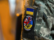 Military jacket with unit patches: Ukrainian flag patch above a round Interflex patch featuring a stylized red creature over a blue-yellow background.