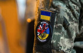 Military jacket with unit patches: Ukrainian flag patch above a round Interflex patch featuring a stylized red creature over a blue-yellow background.