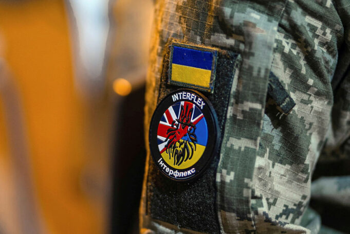 Military jacket with unit patches: Ukrainian flag patch above a round Interflex patch featuring a stylized red creature over a blue-yellow background.