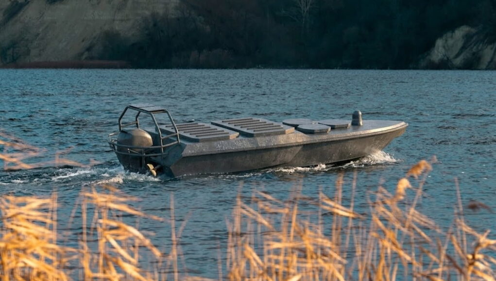 katran-x1-v2 Small aluminum motorboat gliding on a blue lake with a rocky shore in the background, grasses in the foreground.
